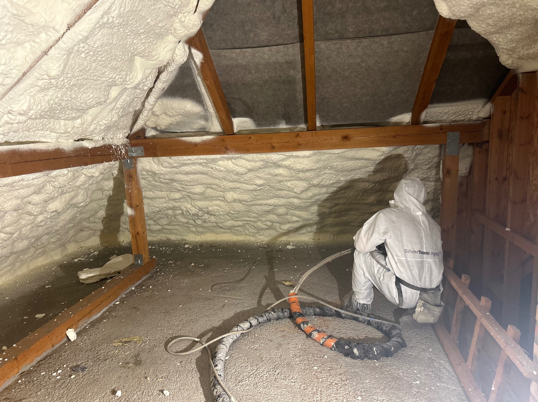 Poddasze w trakcie izolacji pianką PUR, pracownik w kombinezonie ochronnym, widoczne drewniane elementy konstrukcyjne i węże doprowadzające piankę.