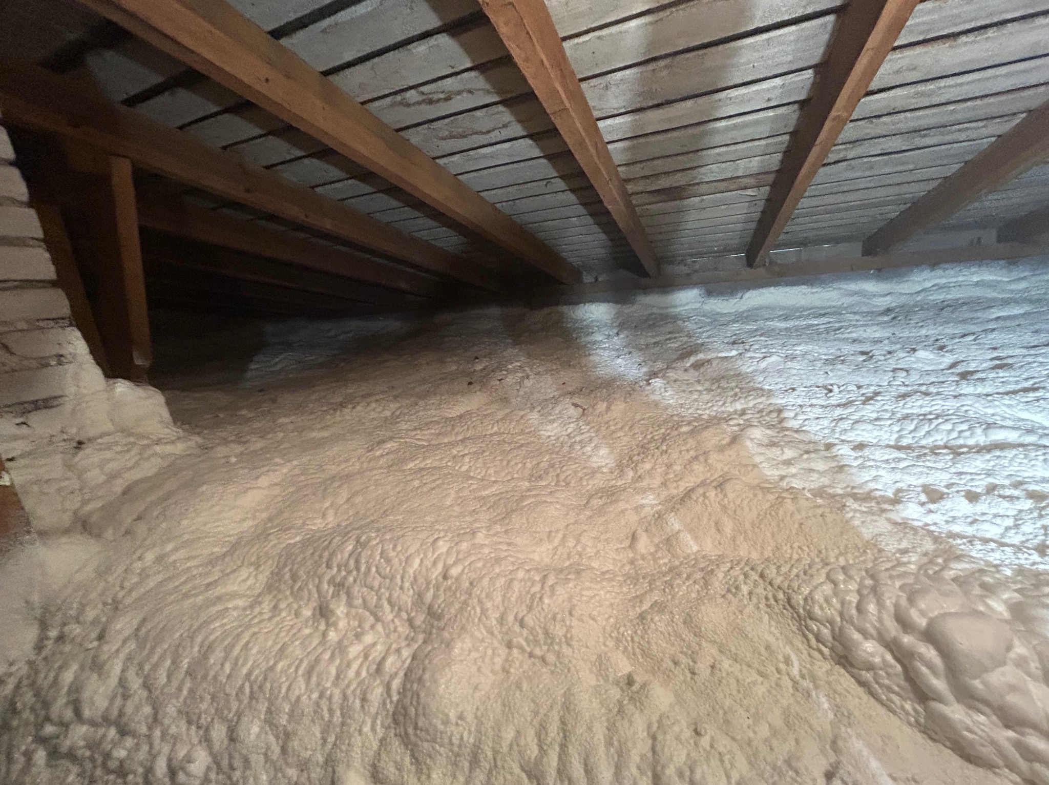 Poddasze z drewnianą konstrukcją pokryte grubą warstwą jasnej pianki izolacyjnej, widoczne fragmenty ceglanej ściany.