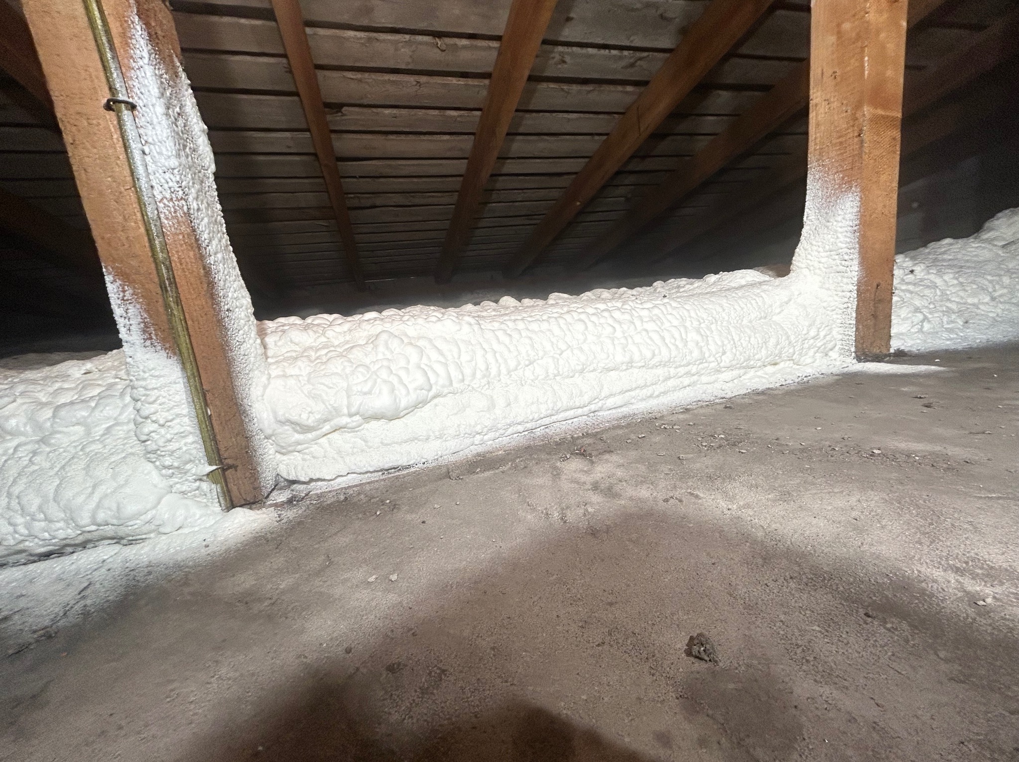 Poddasze ocieplone białą pianką PUR, widoczne drewniane krokwie i fragment betonowej podłogi z drobnymi zabrudzeniami.