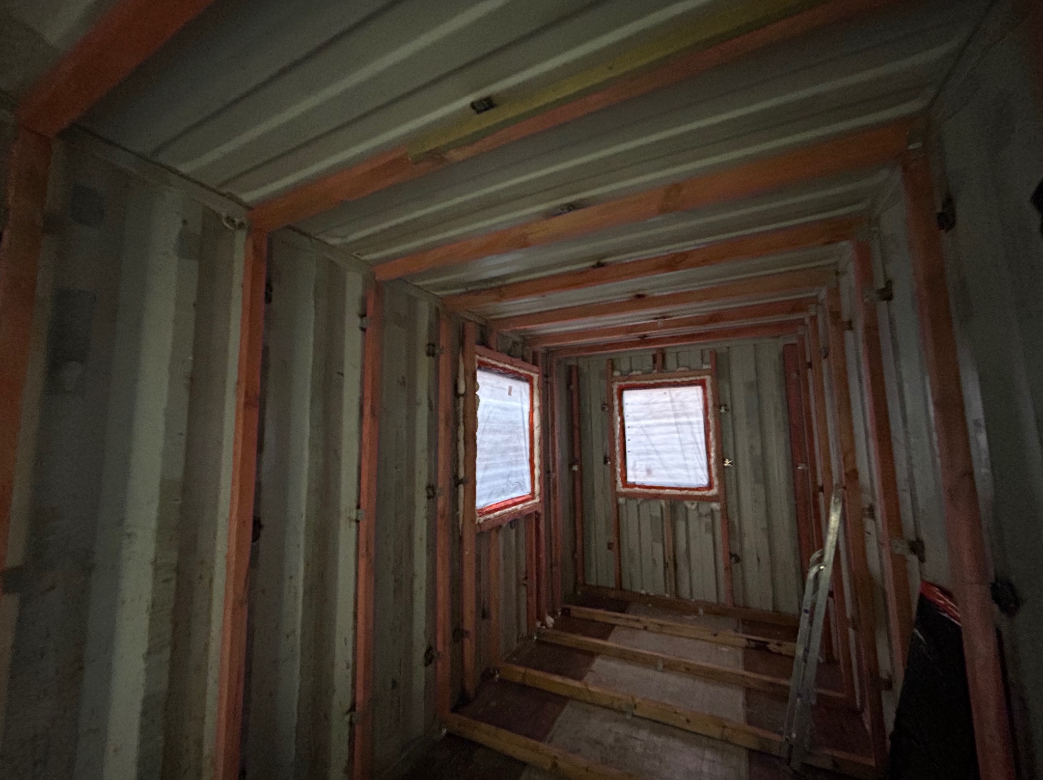 Wnętrze kontenera z widoczną konstrukcją szkieletową ścian i sufitu, przygotowaną pod izolację, z dwoma oknami zabezpieczonymi folią.