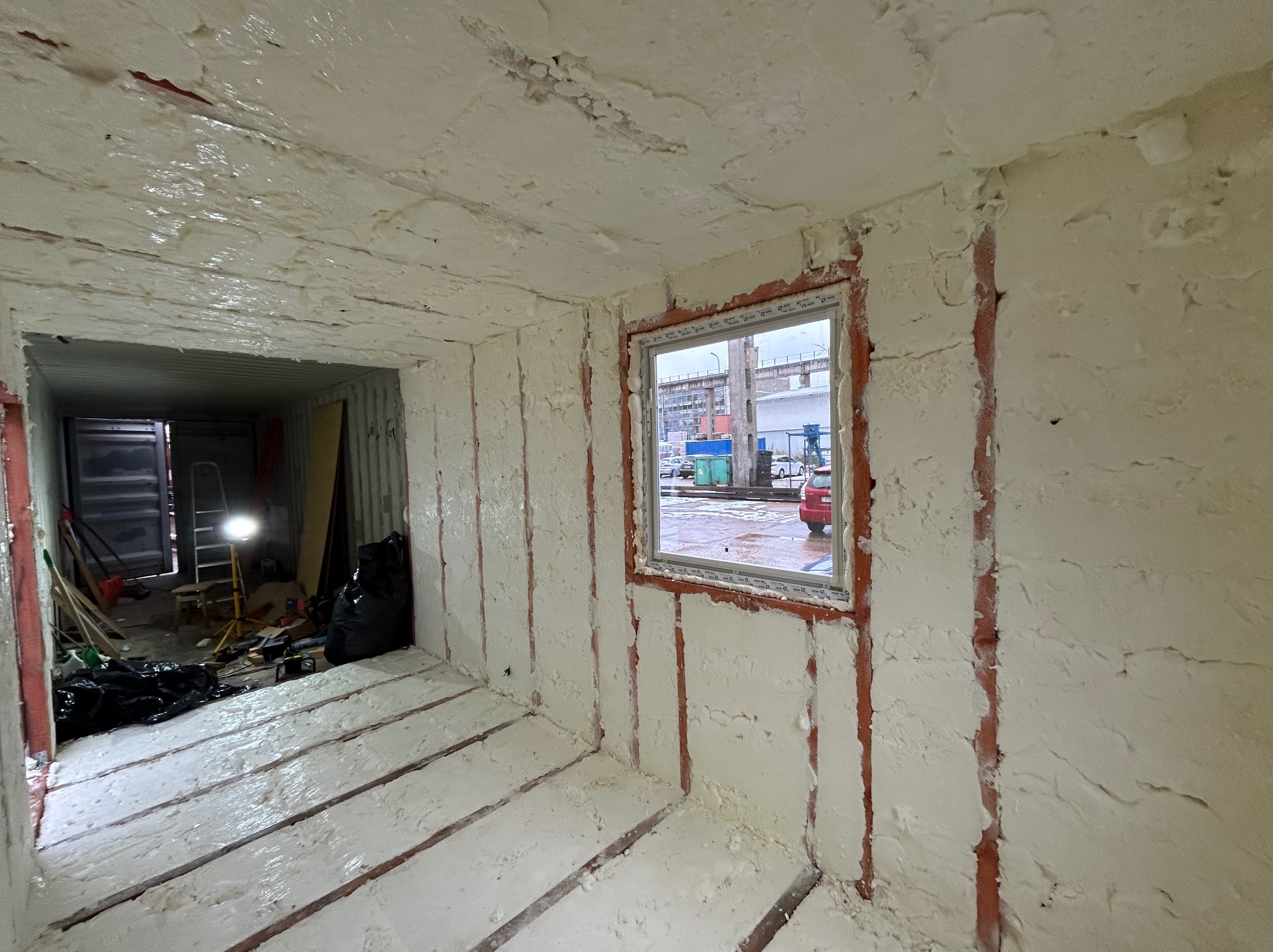 Wnętrze kontenera po natrysku izolacji pianką poliuretanową, widoczne nierównomierne pokrycie pianką na ścianach, suficie i podłodze, zarys konstrukcji kontenera widoczny spod pianki, okno...