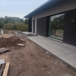 Mateusz Raduj - Nowoczesny dom w trakcie budowy: ułożona kostka brukowa tworzy ścieżkę wzdłuż ściany, widoczne betonowe donice i hałda ziemi na pierwszym planie.