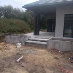 Wykonywanie fundamentów pod taras domu jednorodzinnego w Księżpolu, widoczne betonowe ściany fundamentowe i schody wejściowe, teren wokół w trakcie prac ziemnych.