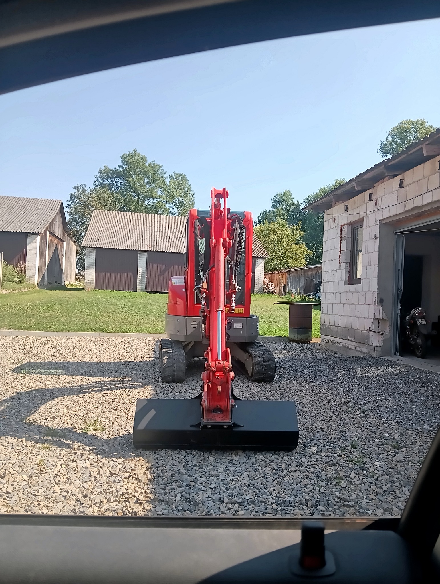 Czerwona mini koparka stoi na żwirowym podwórku, widok z kabiny innego pojazdu, w tle budynki gospodarcze i garaż.