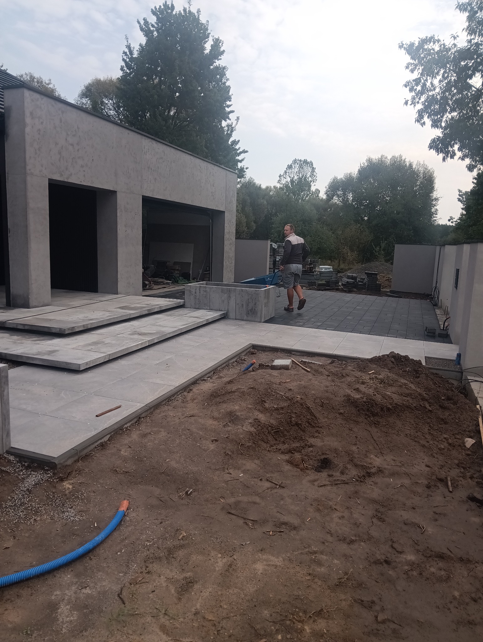 Układanie kostki brukowej na posesji nowoczesnego domu z garażem; widoczne schody z płyt, fragment ułożonej nawierzchni z ciemnej kostki, pracownik z taczką oraz niezagospodarowany teren z rurą...