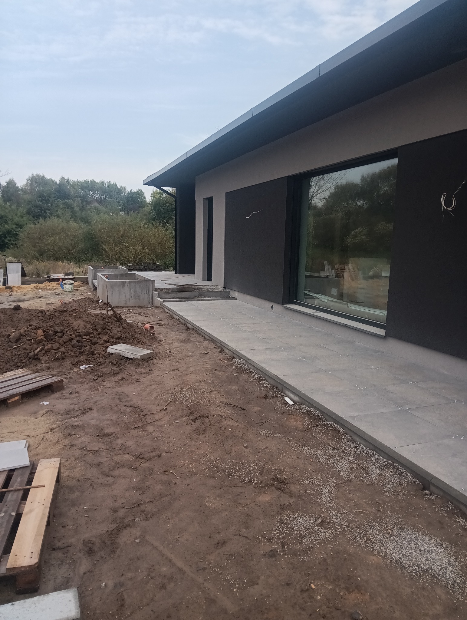 Nowoczesny dom w trakcie budowy: ułożona kostka brukowa tworzy ścieżkę wzdłuż ściany, widoczne betonowe donice i hałda ziemi na pierwszym planie.