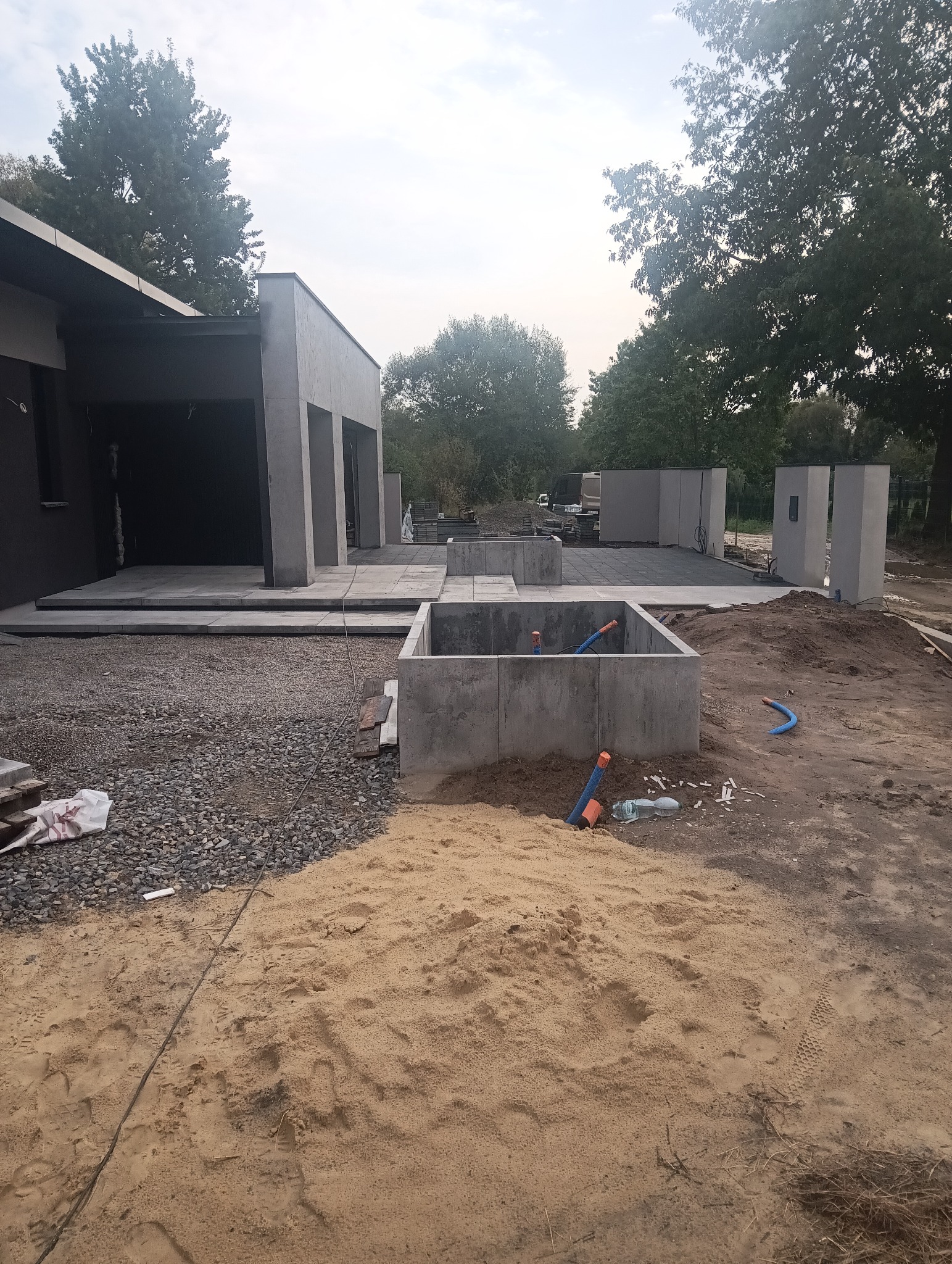 Wykop pod budowę domu z wylanymi betonowymi ścianami, widoczne niebieskie rury instalacyjne wewnątrz wykopu, sterta piasku na pierwszym planie, w tle fragment budynku z szarą elewacją i ułożonymi...