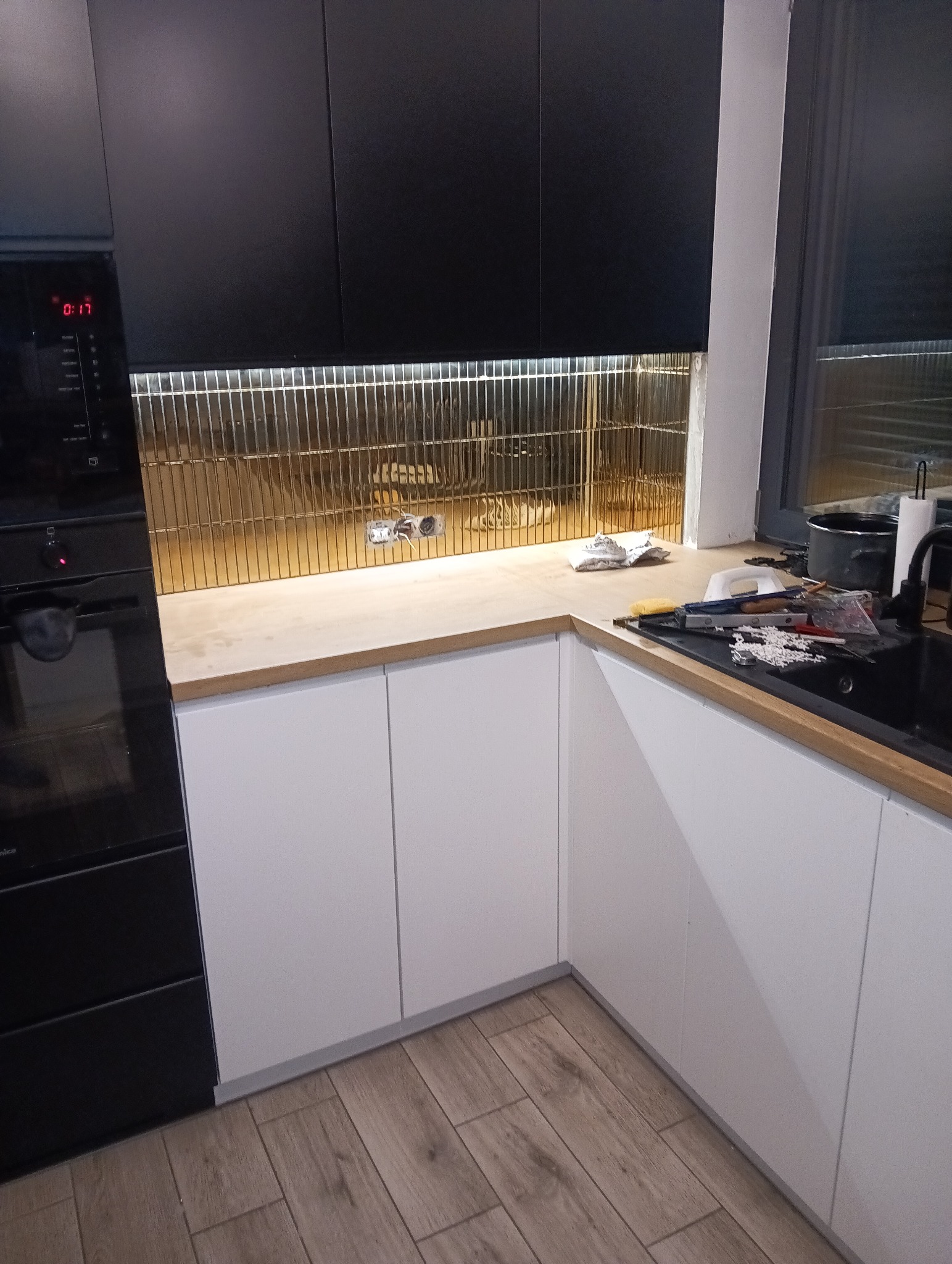 Częściowy widok wnętrza kuchni w trakcie remontu: białe szafki dolne, czarne szafki górne, złota mozaika na ścianie między szafkami, blat z narzędziami i akcesoriami, widoczne gniazdka elektryczne.