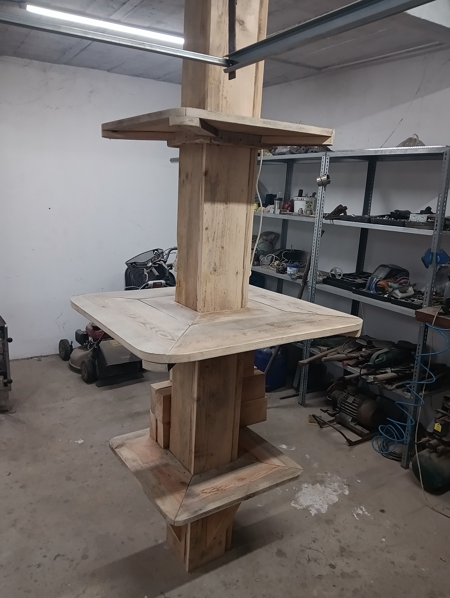 Surowa konstrukcja z drewna, przypominająca szalunek schodkowy lub podstawę pod wylewkę, widoczna w warsztacie z narzędziami i sprzętem w tle.