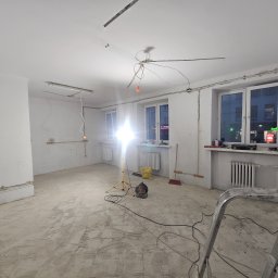 Grupa DEKA - Wnętrze w trakcie remontu: widoczne instalacje elektryczne, przygotowane ściany, podłoga z resztkami kleju, drabina i narzędzia. Oświetlenie robocze.