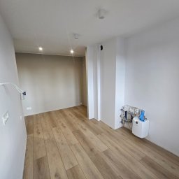 Grupa DEKA - Jasne panele podłogowe w nowym wnętrzu z białymi ścianami. Widoczne gniazdka elektryczne, instalacja grzewcza i oświetlenie sufitowe. Minimalistyczny design.