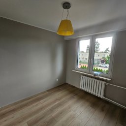 Grupa DEKA - Pokój po remoncie: drewnopodobne panele, szare ściany, biały grzejnik pod oknem z widokiem na zieleń. Żółta lampa wisząca dodaje koloru.