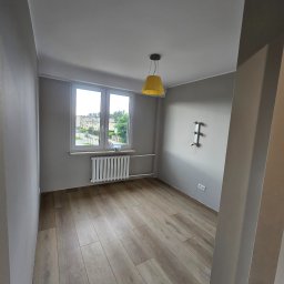 Grupa DEKA - Jasne panele podłogowe w pokoju z szarymi ścianami i oknem. Żółta lampa wisząca dodaje koloru. Widoczny grzejnik i instalacja elektryczna.