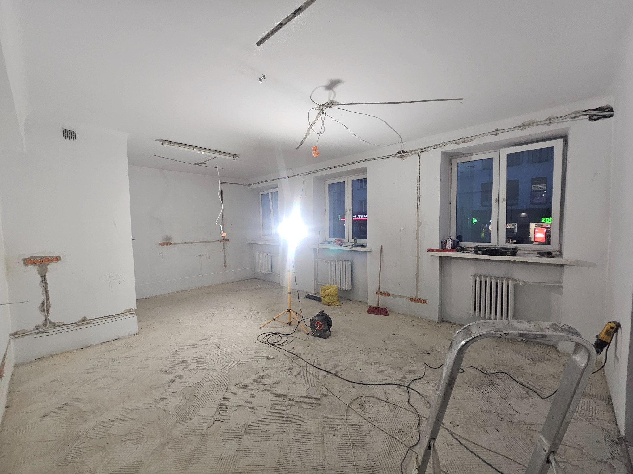 Wnętrze w trakcie remontu: widoczne instalacje elektryczne, przygotowane ściany, podłoga z resztkami kleju, drabina i narzędzia. Oświetlenie robocze.