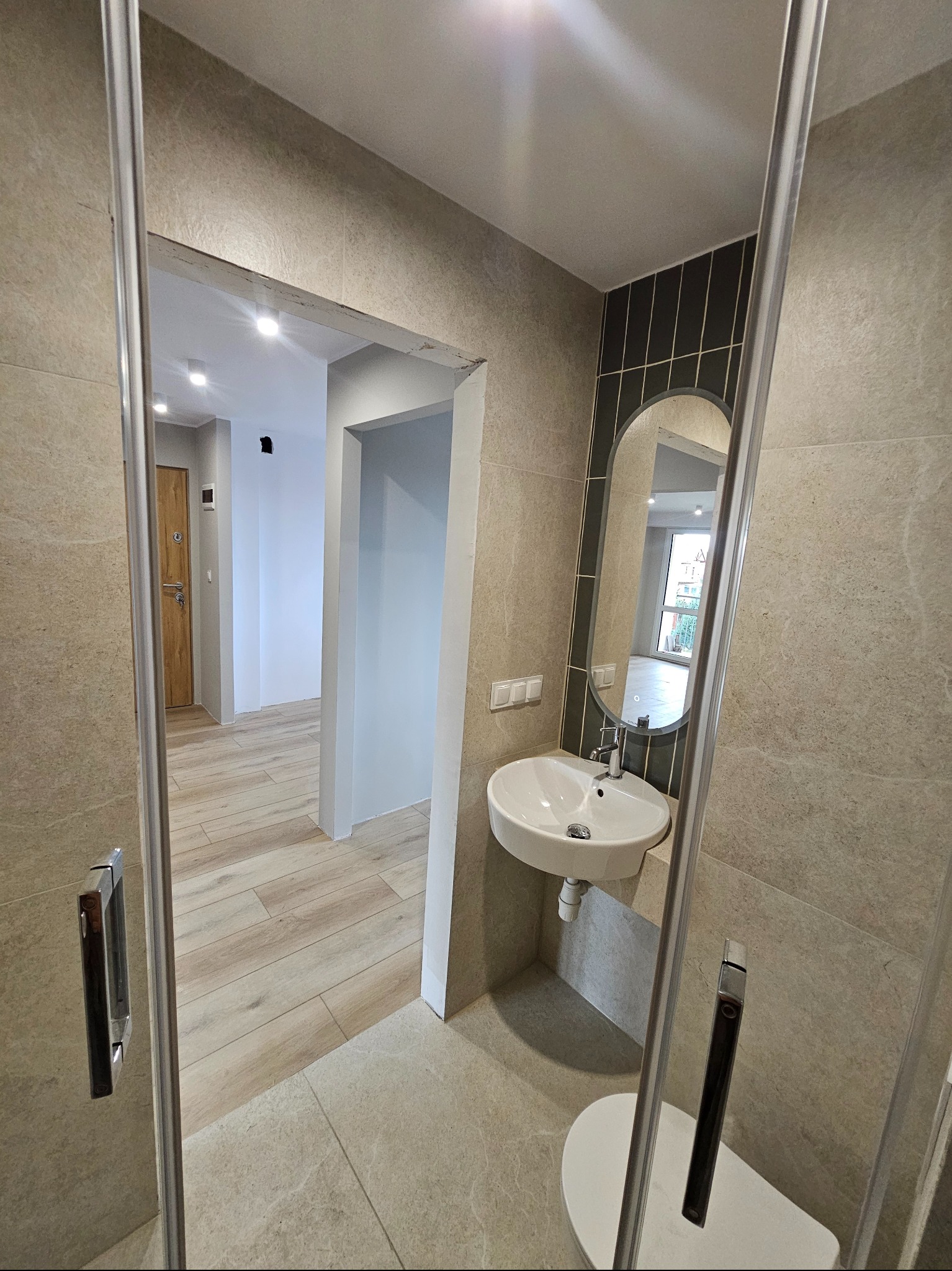 Nowoczesna łazienka z kabiną prysznicową typu walk-in, owalnym lustrem nad umywalką i beżowymi płytkami. Widok na korytarz z drewnianą podłogą i drzwiami.