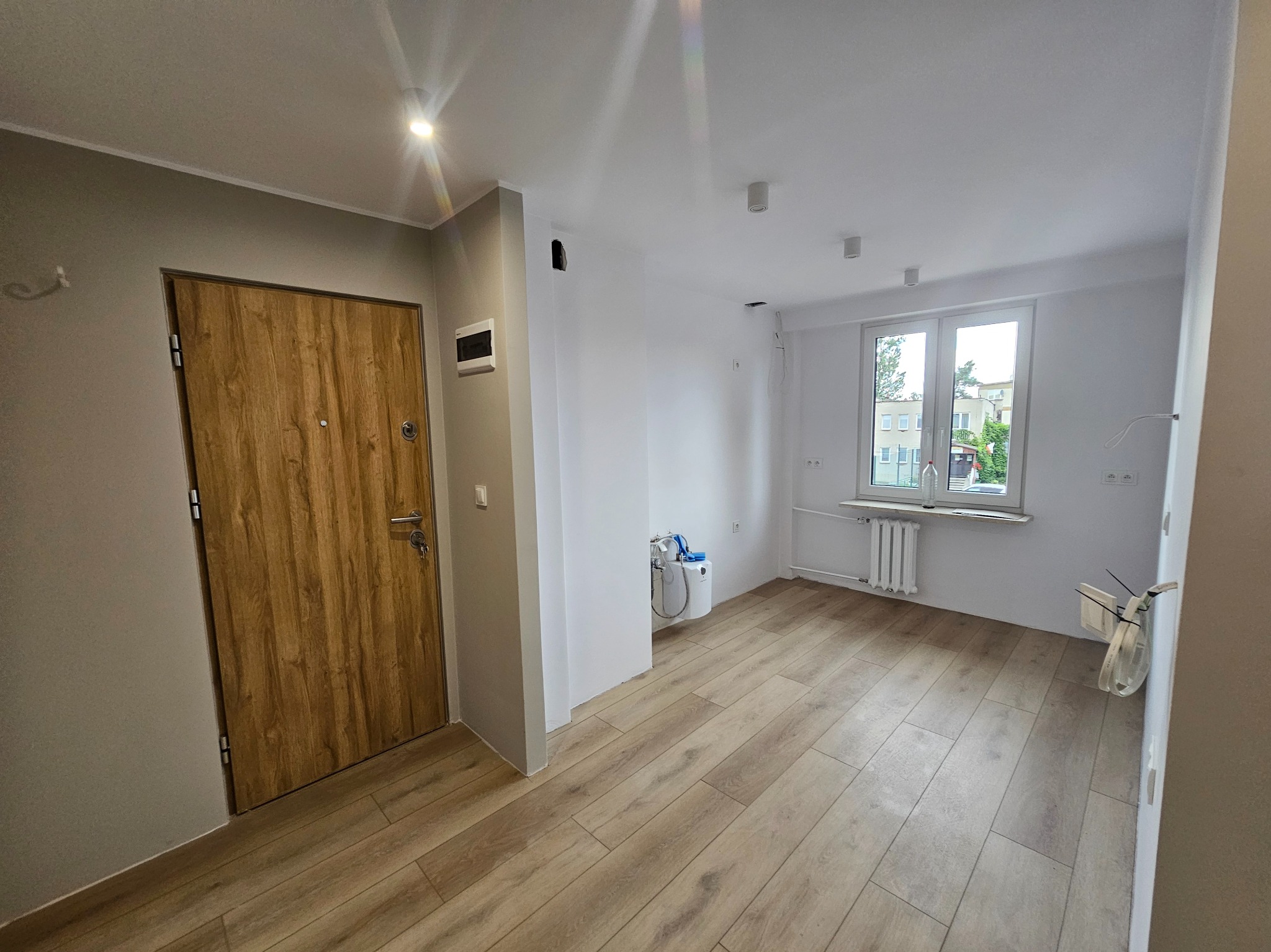 Jasne wnętrze po remoncie: drewnopodobne panele, białe ściany, okno z widokiem na zieleń. Nowe drzwi wejściowe z drewnianym wykończeniem. Minimalistyczne oświetlenie.