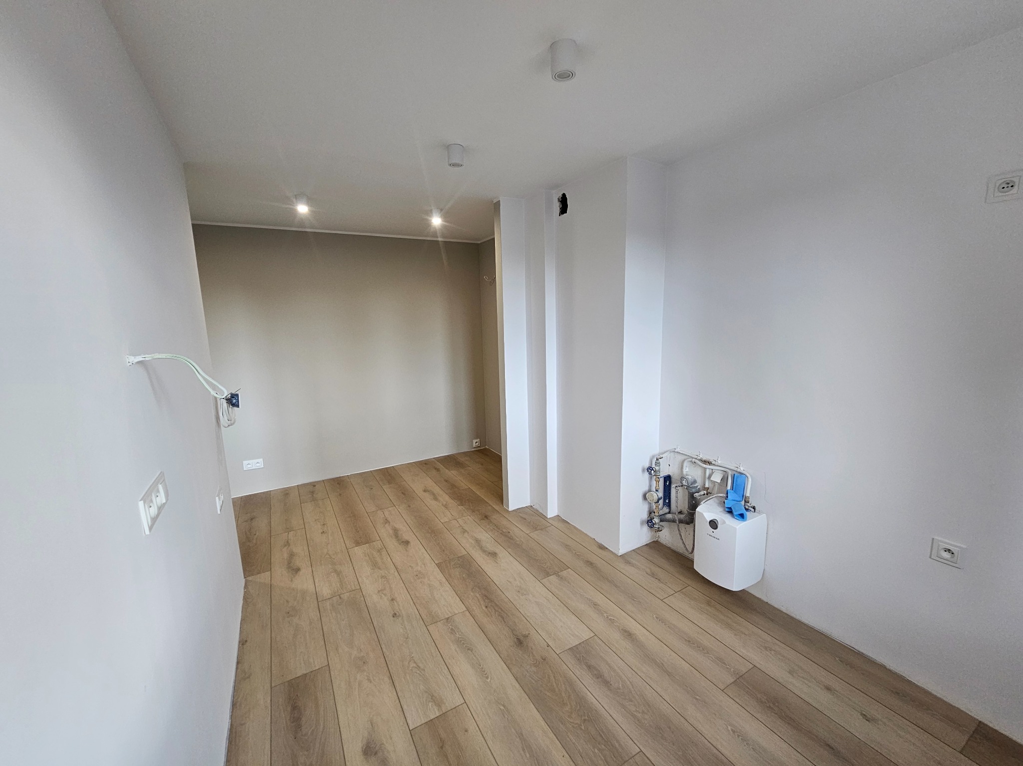 Jasne panele podłogowe w nowym wnętrzu z białymi ścianami. Widoczne gniazdka elektryczne, instalacja grzewcza i oświetlenie sufitowe. Minimalistyczny design.