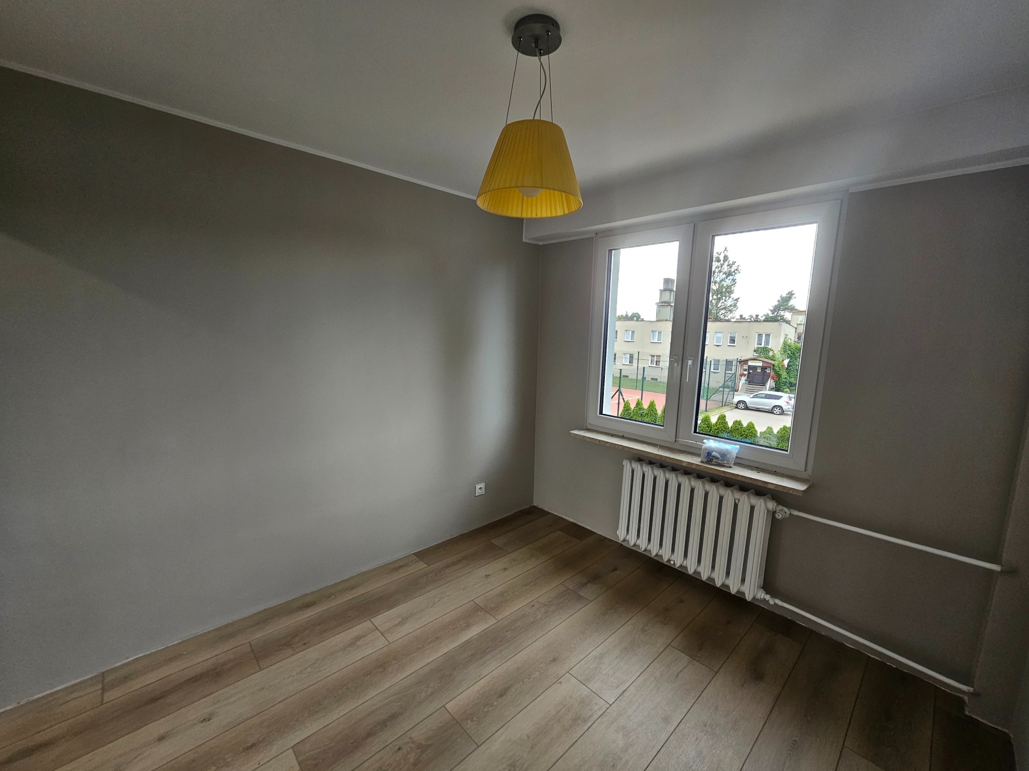 Pokój po remoncie: drewnopodobne panele, szare ściany, biały grzejnik pod oknem z widokiem na zieleń. Żółta lampa wisząca dodaje koloru.
