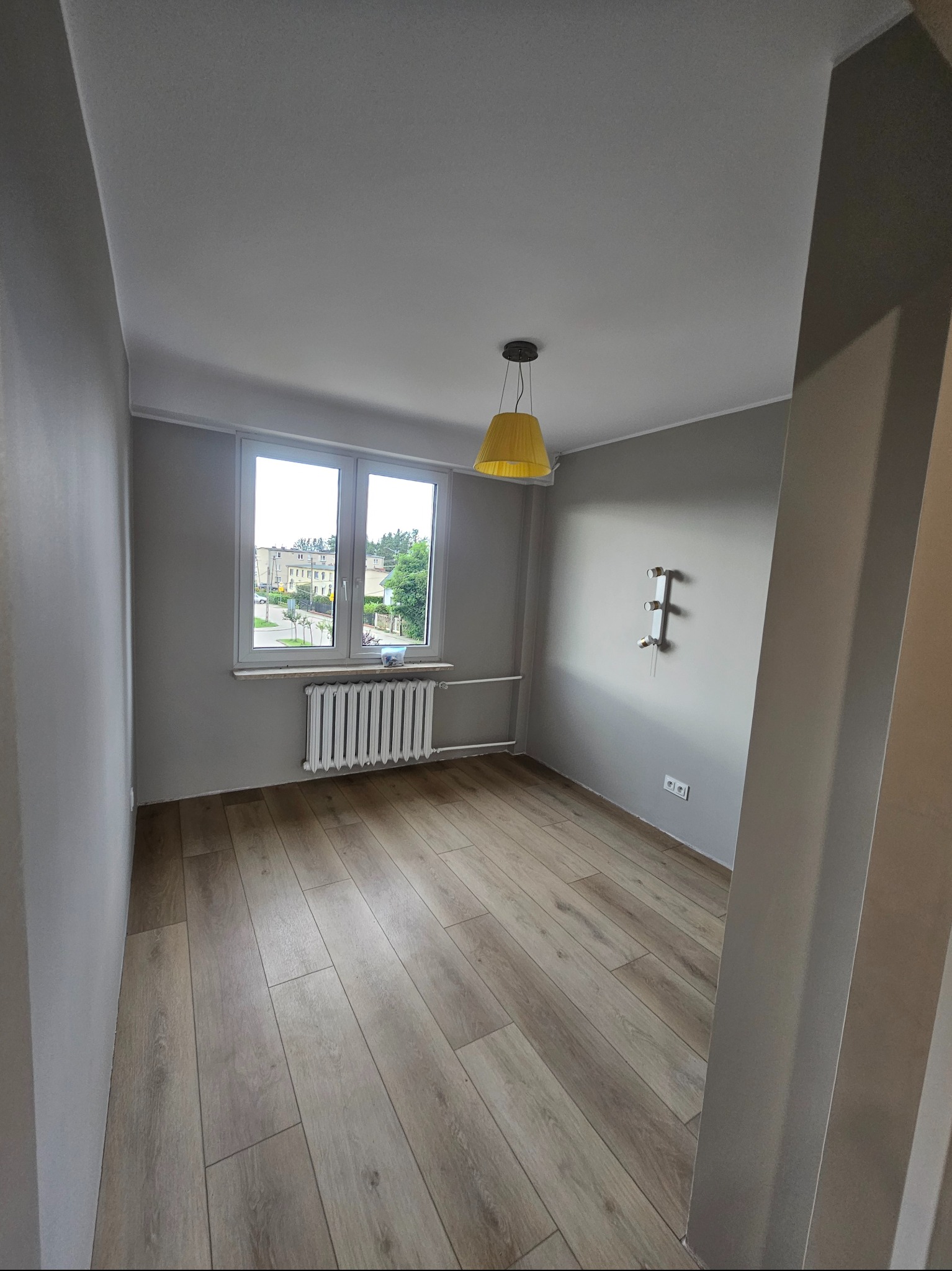 Jasne panele podłogowe w pokoju z szarymi ścianami i oknem. Żółta lampa wisząca dodaje koloru. Widoczny grzejnik i instalacja elektryczna.