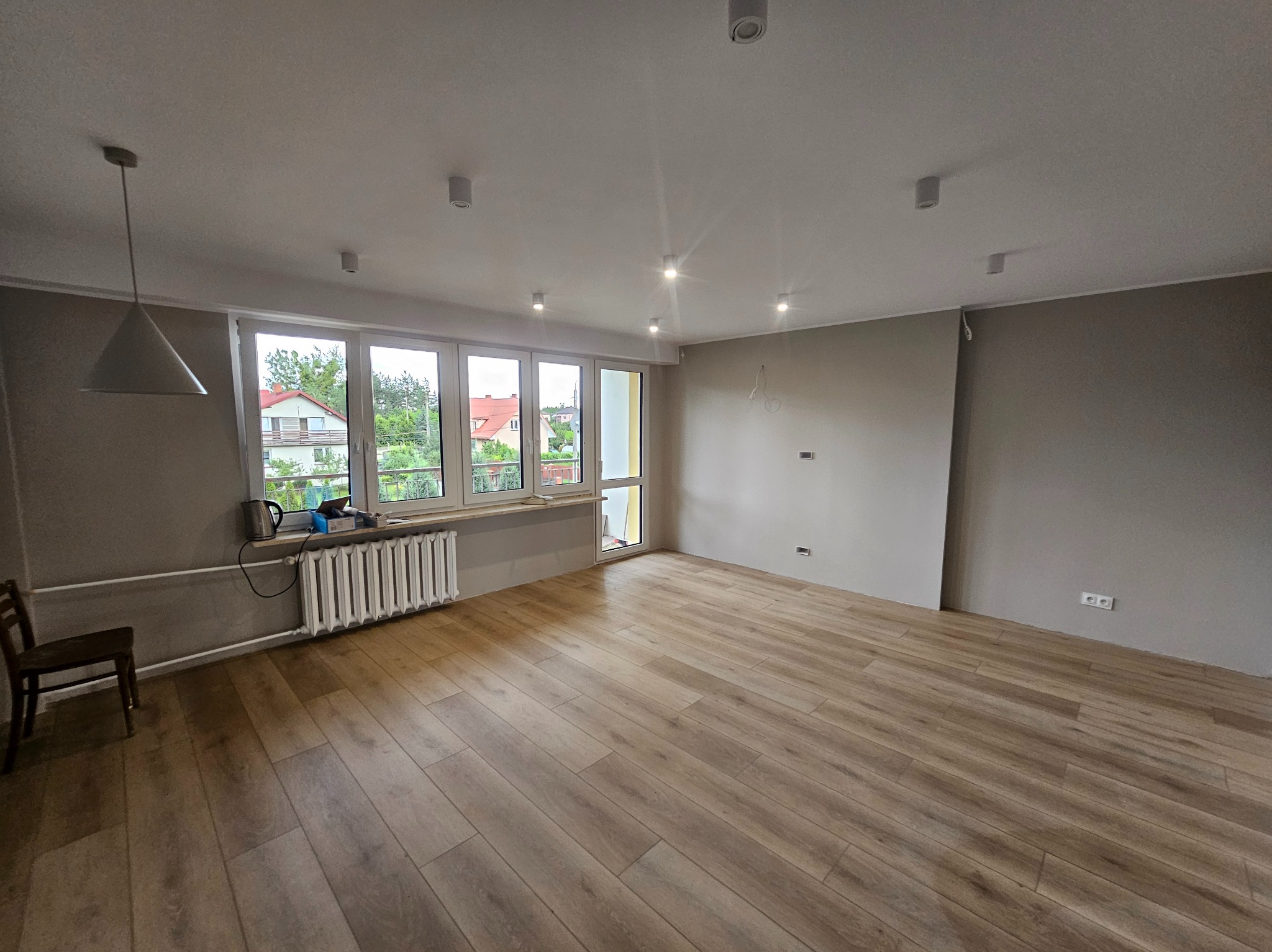 Jasne panele podłogowe w pustym pokoju z grzejnikiem pod oknem. Świeżo pomalowane ściany w odcieniach szarości. Widoczne lampy sufitowe i pojedyncze krzesło.
