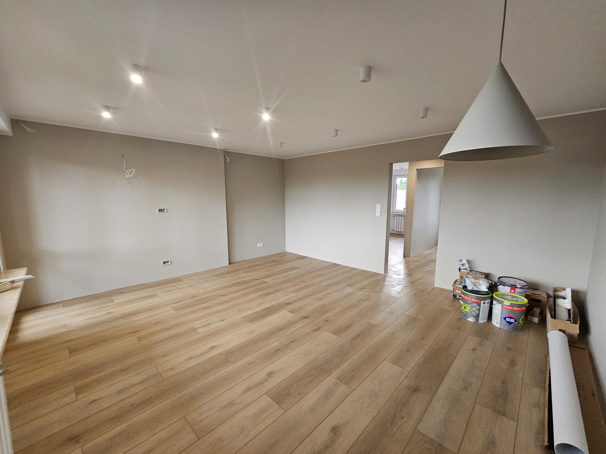 Jasne panele podłogowe w przestronnym, odświeżonym pokoju z szarymi ścianami i nowoczesnym oświetleniem. Widoczne puszki z farbą i lampa w kształcie stożka.