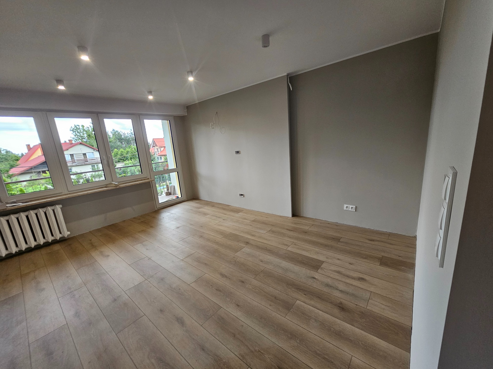 Jasne panele podłogowe w nowym pokoju z grzejnikiem pod oknem i szarą ścianą. Widoczne gniazdka elektryczne i punkty świetlne na suficie. Świeżo po remoncie.