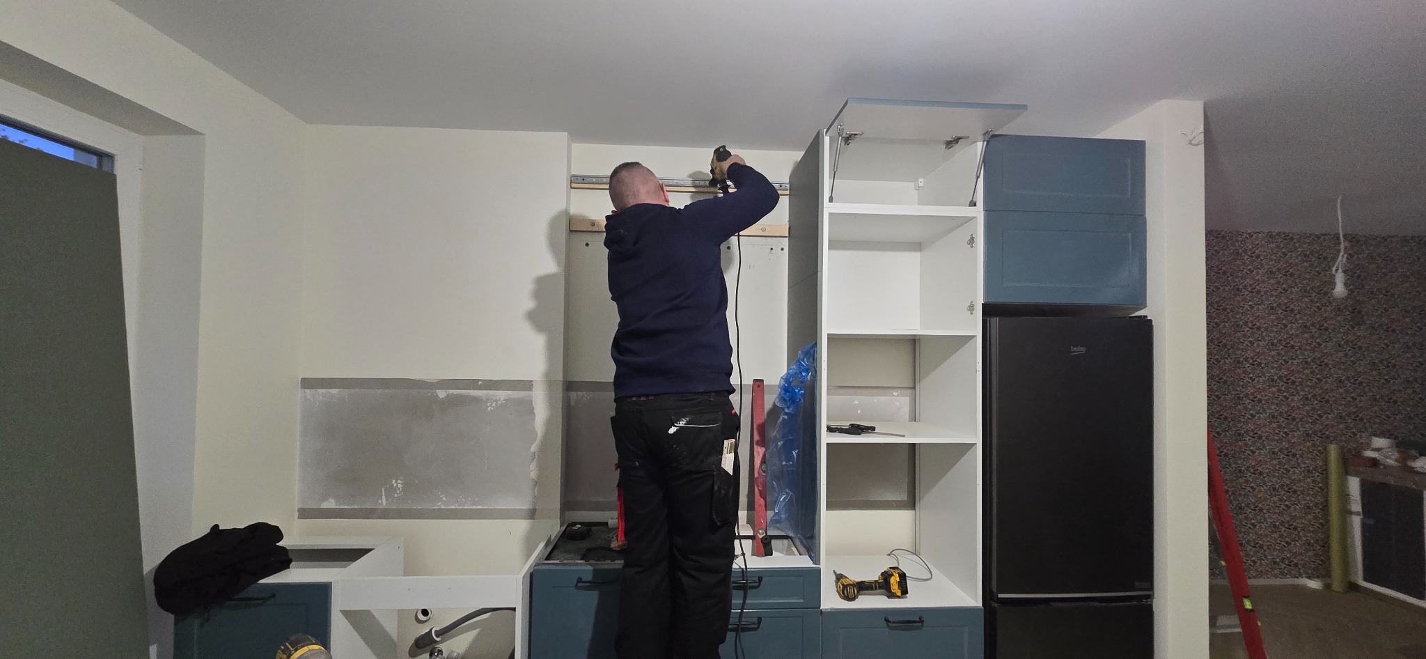 Monterażysta w trakcie montażu szafki kuchennej z niebieskimi frontami. Widoczne narzędzia i elementy instalacji. Ściana przygotowana pod zabudowę.