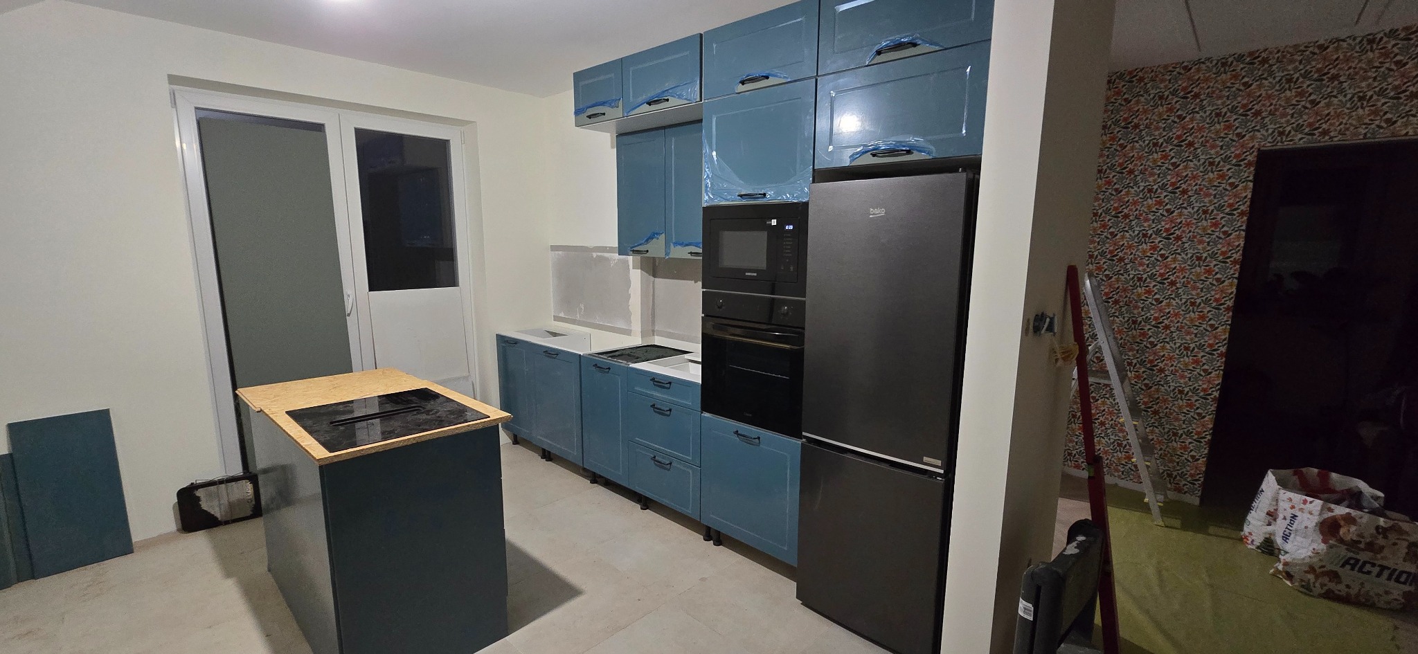 Niebieska zabudowa kuchenna z wyspą i czarnym sprzętem AGD, częściowo jeszcze zabezpieczona folią. Widoczne przygotowania do wykończenia ścian.