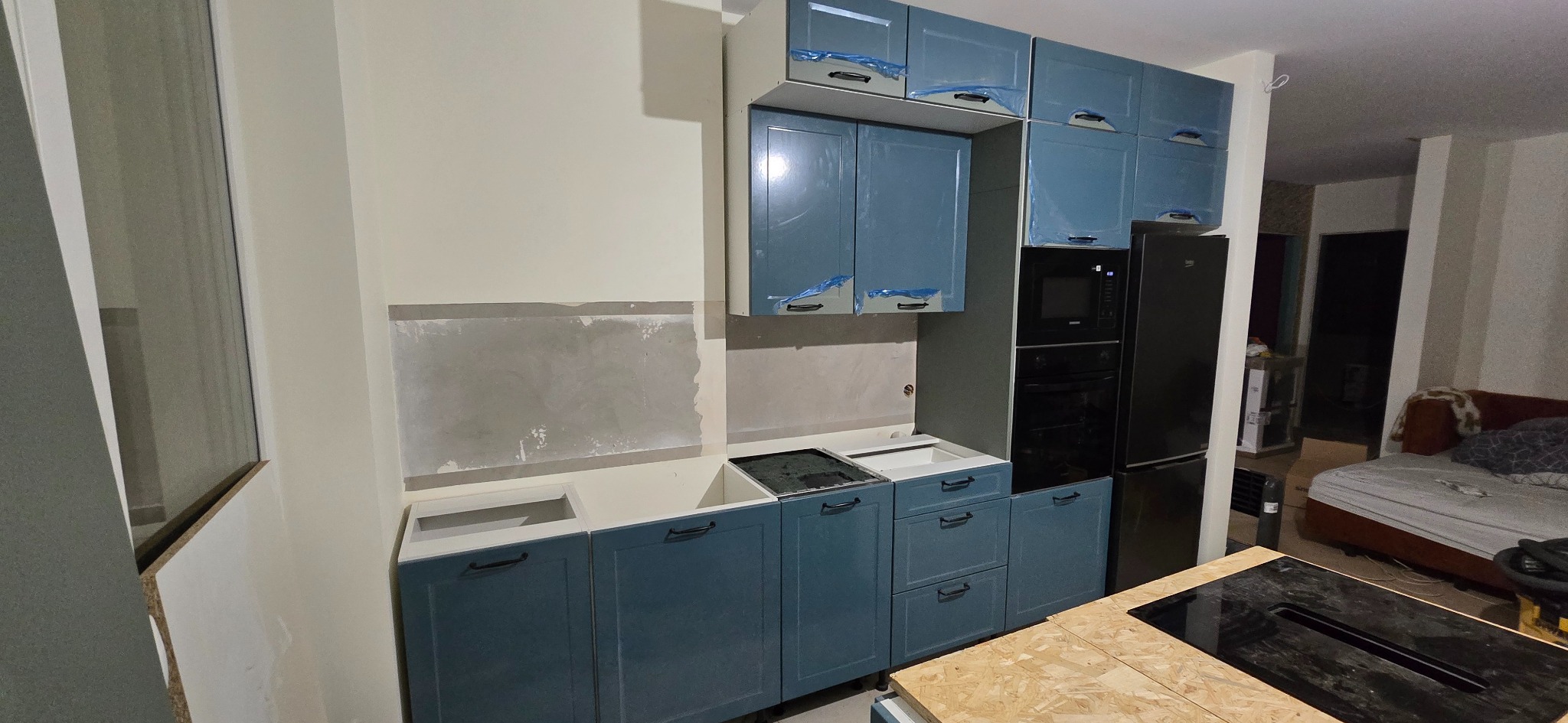 Niebieska zabudowa kuchenna w trakcie montażu, z folią ochronną na frontach. Ściana nad blatem przygotowana pod płytki. Widoczny sprzęt AGD i fragment wyspy kuchennej.