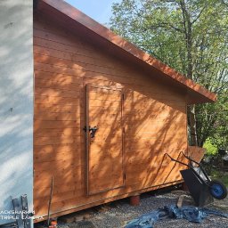 Jonasz Henszel - Świeżo wybudowana, drewniana szopa ogrodowa z dwuskrzydłowymi drzwiami, widoczna z boku, z taczką i narzędziami w pobliżu, na tle zieleni.