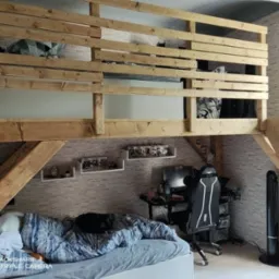 Drewniana antresola z łóżkiem na górze i przestrzenią do pracy z biurkiem i krzesłem gamingowym na dole, pokój z tapetą imitującą cegłę i półkami na ścianie.