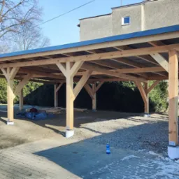 Drewniana konstrukcja z czarnym dachem w trakcie budowy, widoczne słupy na betonowych podstawach, materiały budowlane i butelka wody na brukowanym podłożu, dom w tle.