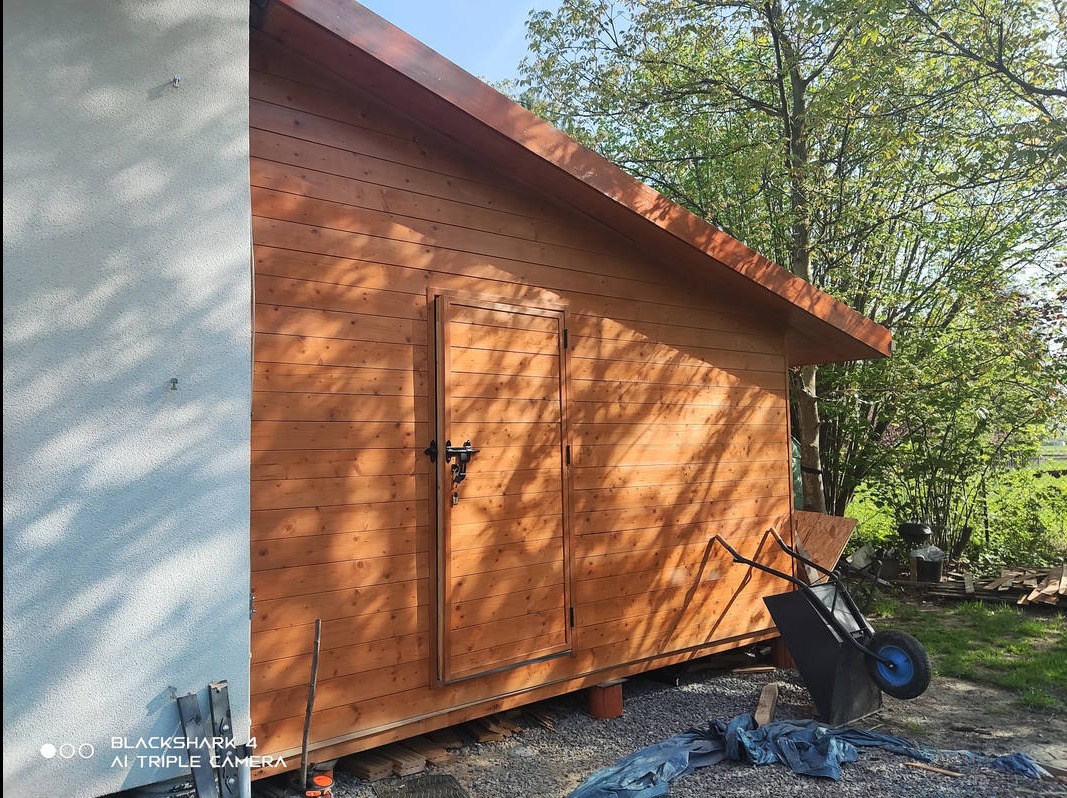 Świeżo wybudowana, drewniana szopa ogrodowa z dwuskrzydłowymi drzwiami, widoczna z boku, z taczką i narzędziami w pobliżu, na tle zieleni.