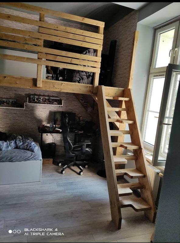 Drewniana antresola z drabinką w pokoju, widok na biurko i łóżko, naturalne drewno, schody z wycięciami na stopy.
