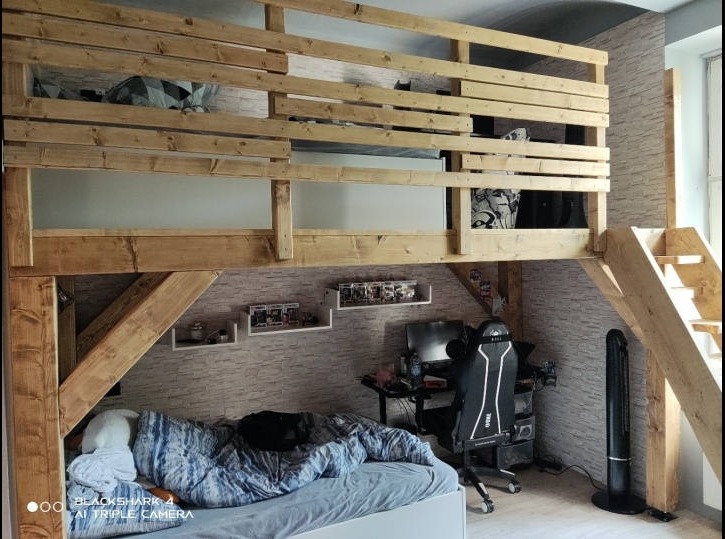 Drewniana antresola z łóżkiem na górze i przestrzenią do pracy z biurkiem i krzesłem gamingowym na dole, pokój z tapetą imitującą cegłę i półkami na ścianie.