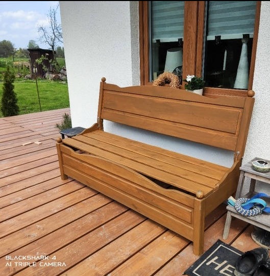 Drewniana skrzynia-ławka z oparciem na tarasie, widoczne uszkodzone deski siedziska, obok okno z dekoracjami, na podłodze wycieraczka z napisem 'HE'.