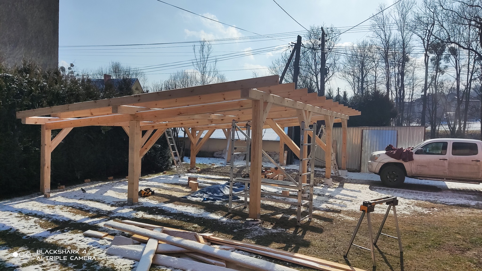 Konstrukcja z surowego drewna w trakcie budowy, prawdopodobnie zadaszenie lub wiata, widoczne drabiny i narzędzia, lekko zaśnieżony teren, samochód w tle.
