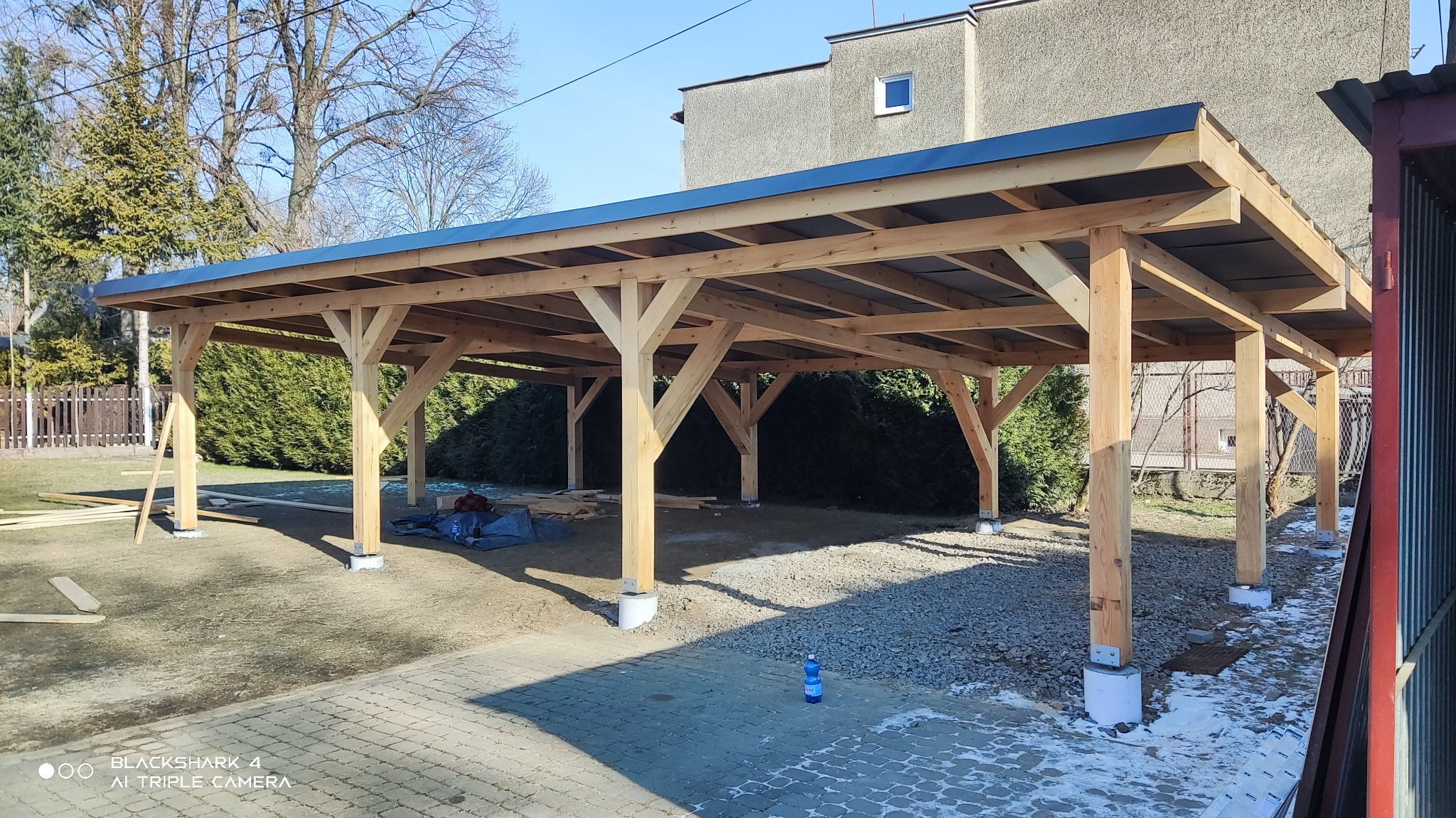 Drewniana konstrukcja z czarnym dachem w trakcie budowy, widoczne słupy na betonowych podstawach, materiały budowlane i butelka wody na brukowanym podłożu, dom w tle.