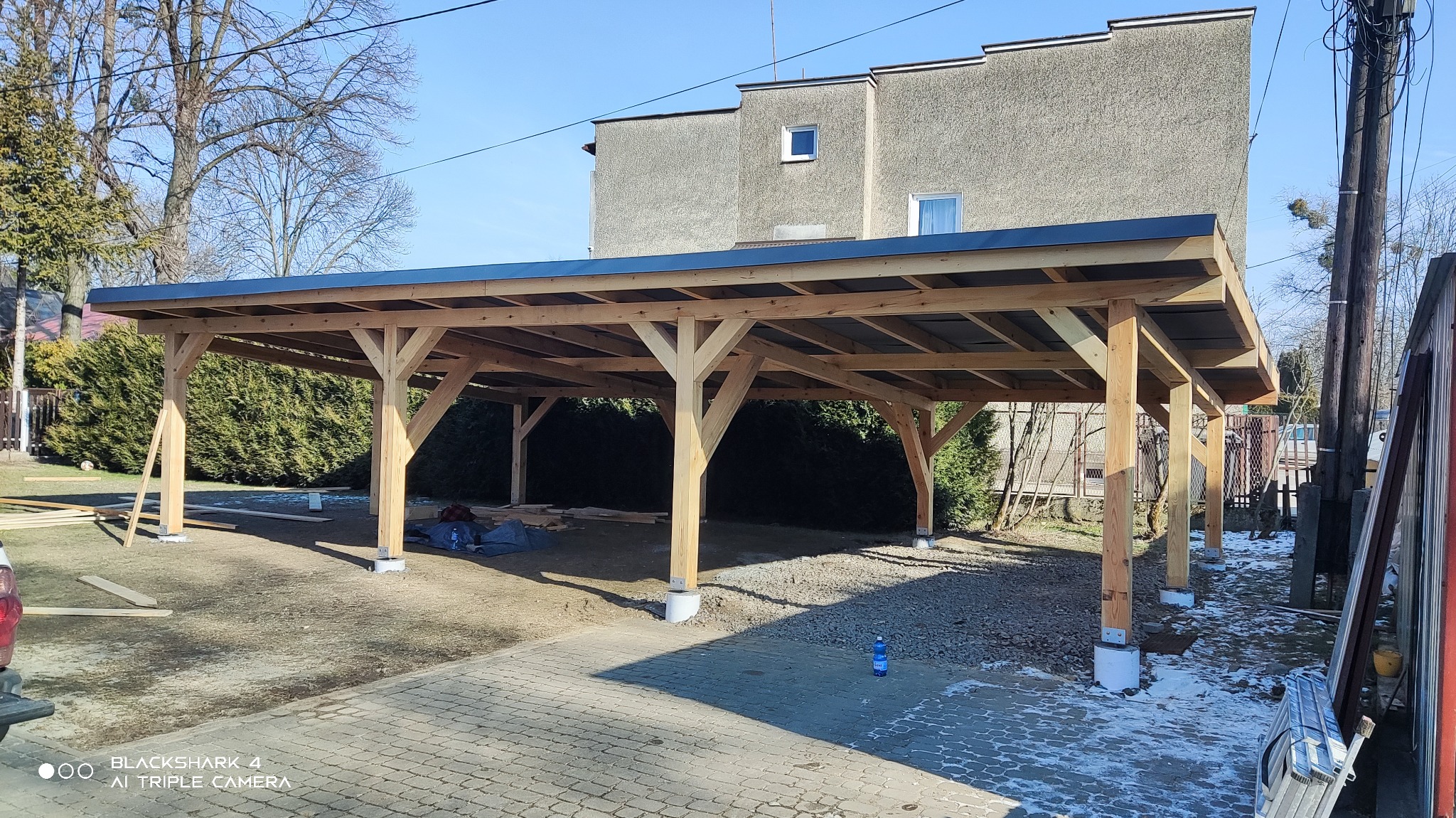 Drewniana konstrukcja z czarnym dachem, prawdopodobnie zadaszenie tarasu lub wiata garażowa, w trakcie budowy, widoczne drewniane elementy i betonowe podstawy słupów.