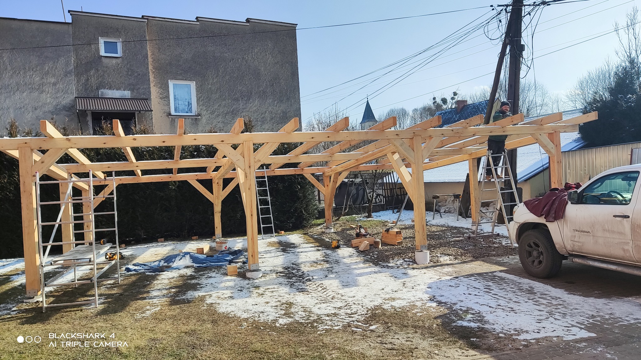 Konstrukcja z jasnego drewna w trakcie montażu, prawdopodobnie zadaszenie tarasu lub pergola, widoczna osoba na drabinie, narzędzia pracy, częściowo ośnieżony teren.