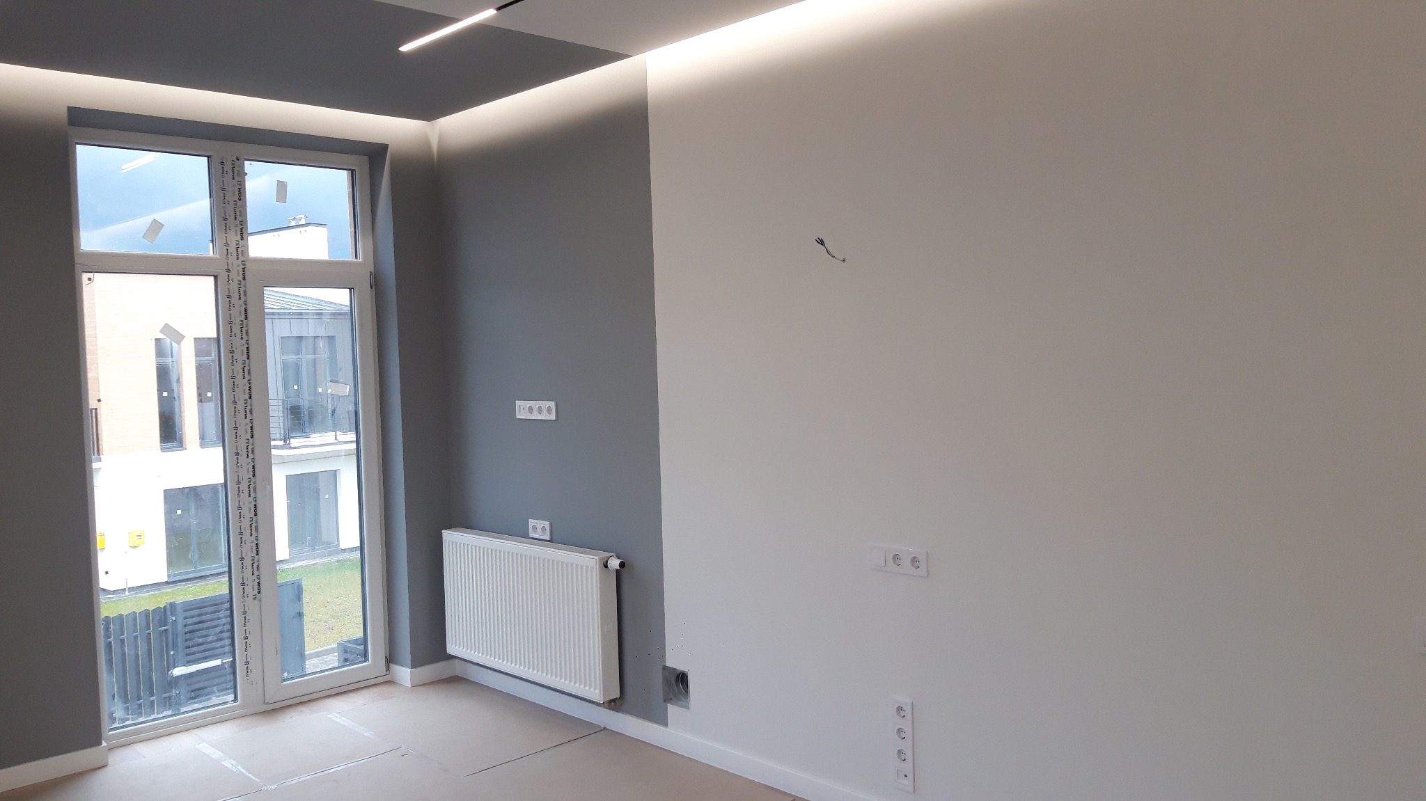 Pokój po malowaniu, z szarą ścianą i białą ścianą, białym grzejnikiem pod oknem i gniazdkami elektrycznymi, widok na zewnątrz przez okno.