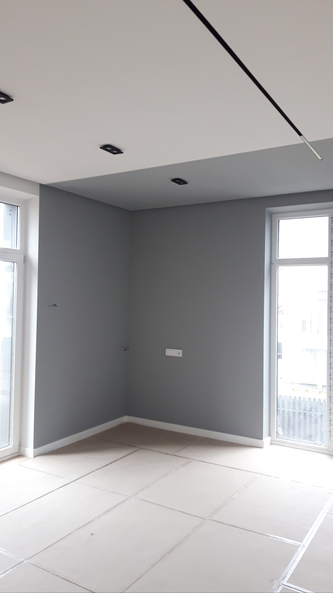 Szare ściany w pustym pokoju z białymi oknami i przygotowanym podłożem pod panele, widoczne czarne, nowoczesne lampy sufitowe.
