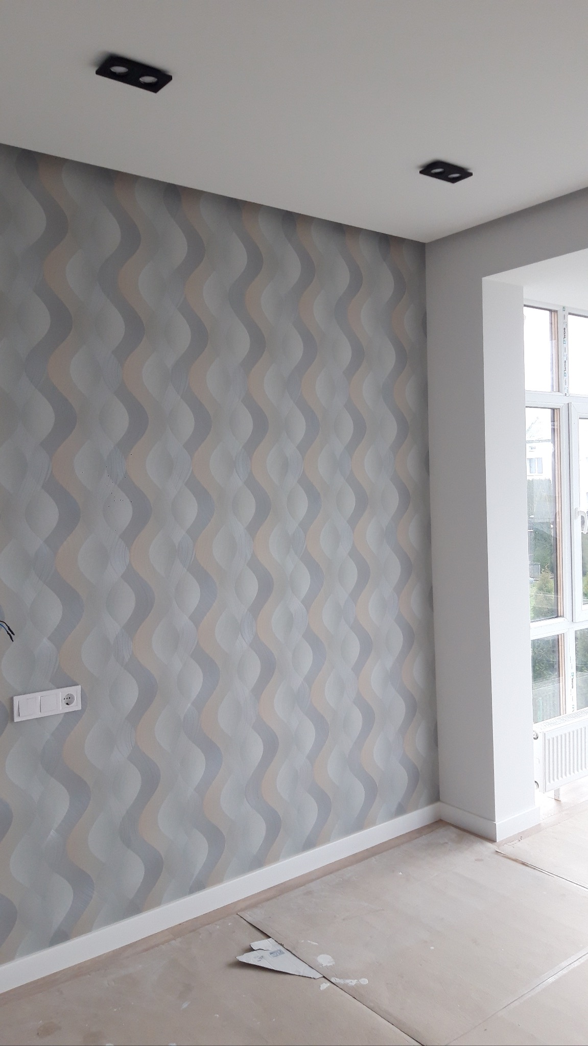 Wnętrze pokoju po tapetowaniu. Ściana pokryta tapetą w odcieniach szarości i beżu z falistym wzorem. Widoczne gniazdko, listwa przypodłogowa i fragment okna.
