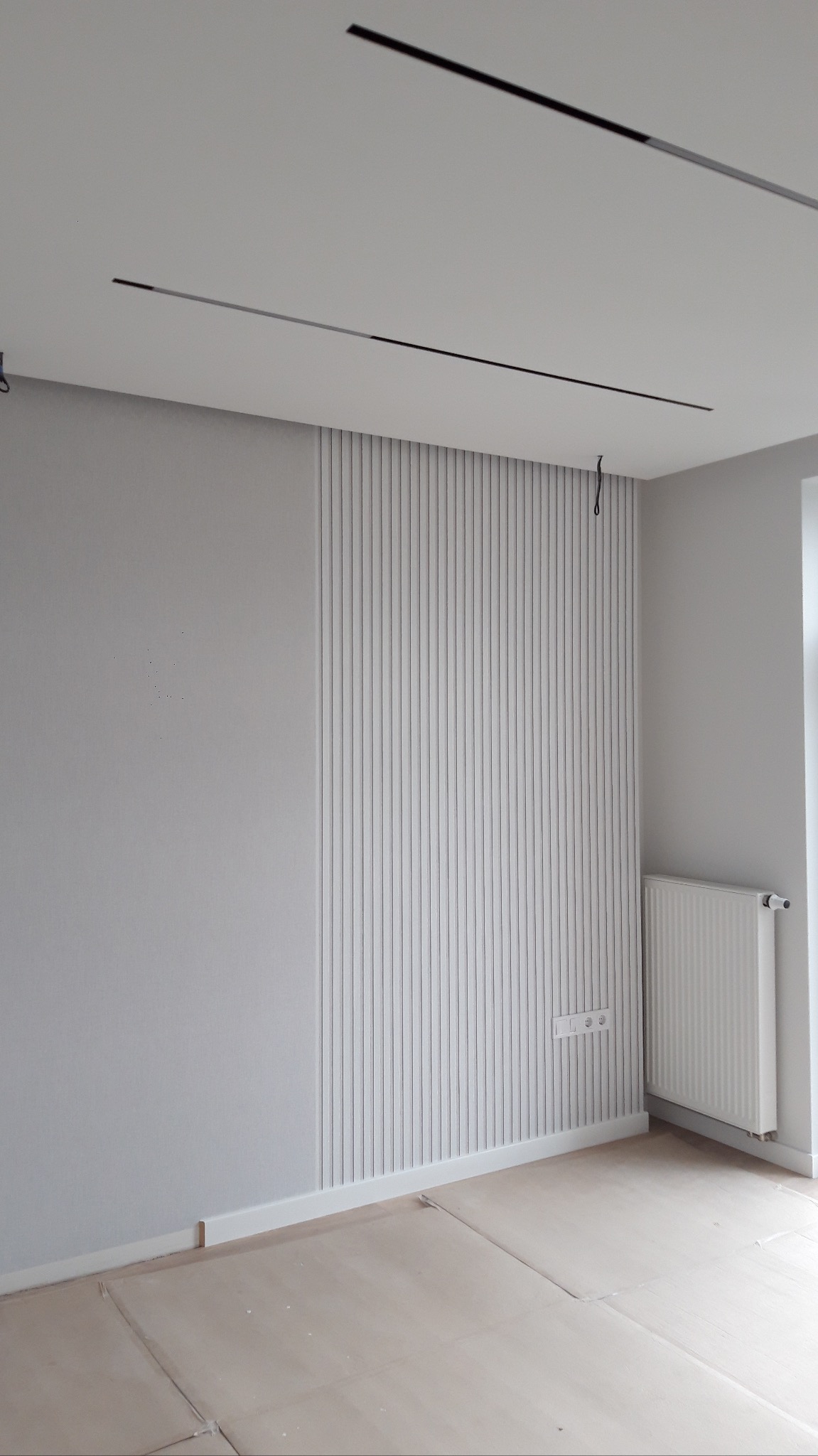 Szara ściana z dekoracyjnymi, pionowymi panelami. Widoczne listwy przypodłogowe i minimalistyczne oświetlenie sufitowe. Na podłodze zabezpieczenie.