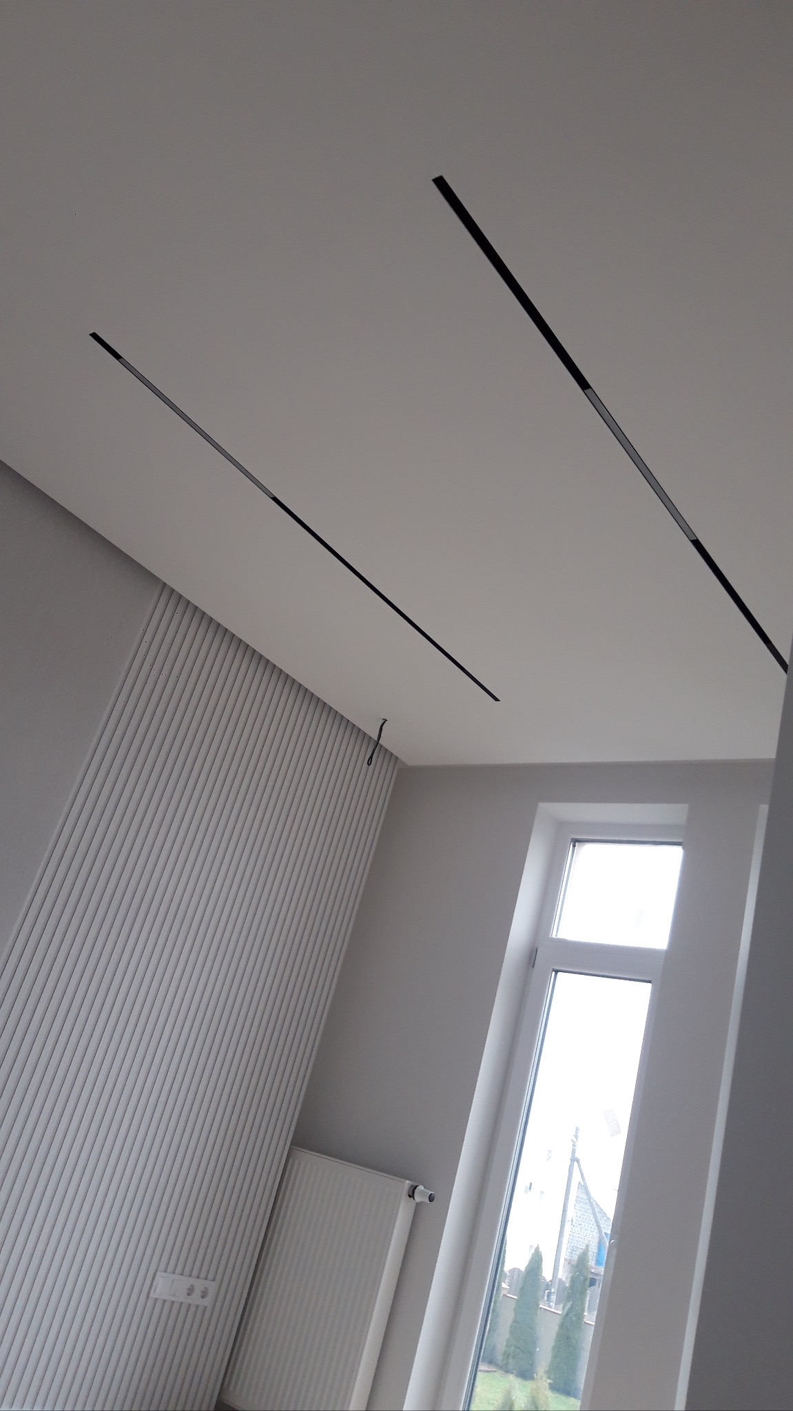 Nowoczesne wykończenie wnętrza: biały sufit z czarnymi, liniowymi lampami LED, panel ścienny z pionowymi żłobieniami, okno. Minimalistyczny design.