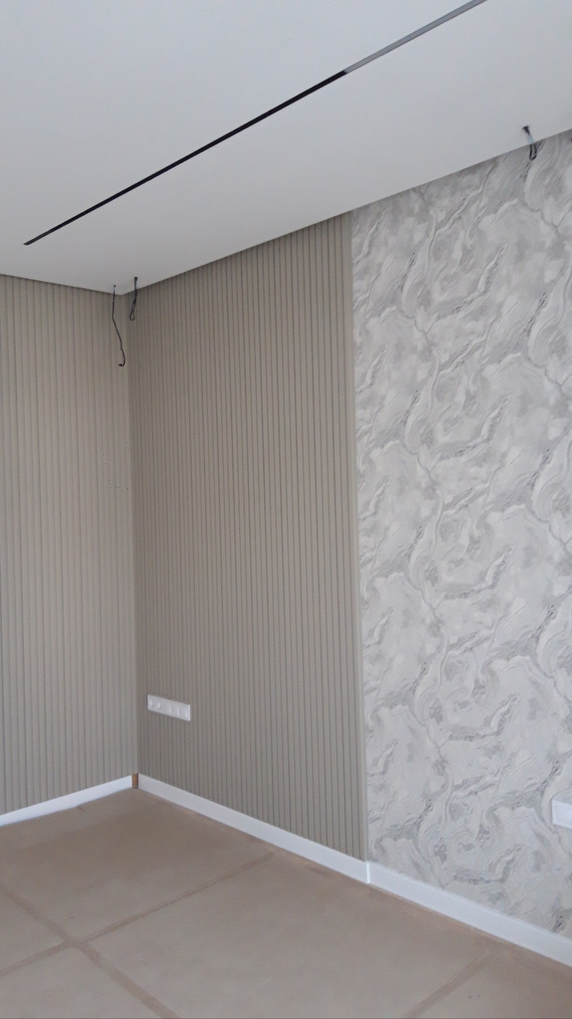 Wnętrze pokoju po remoncie: ściana z beżowymi panelami pionowymi i marmurową tapetą, biały sufit z czarnym paskiem oświetleniowym, jasna podłoga z płytkami.