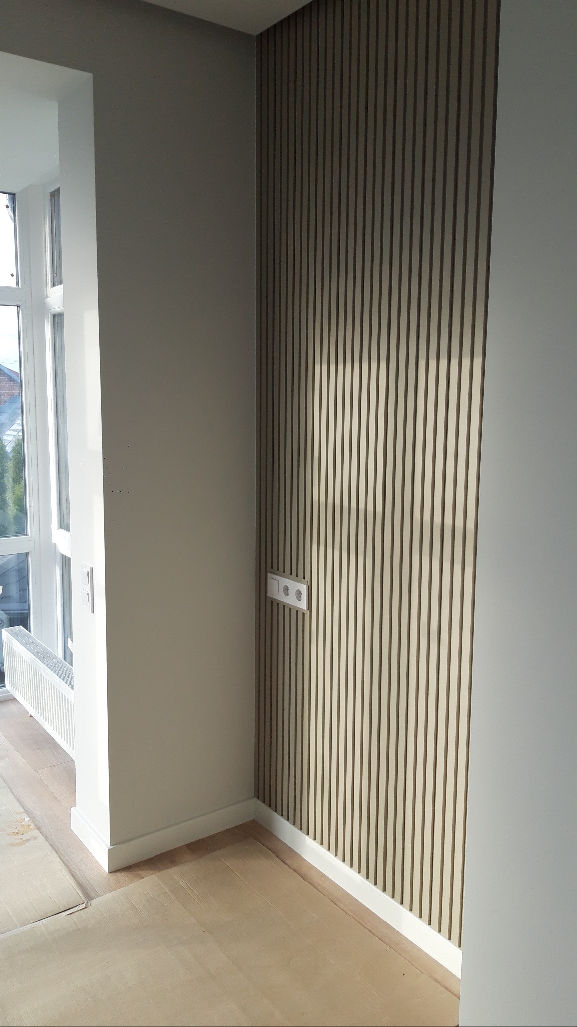 Nowoczesne wykończenie wnętrza: ściana z drewnianych paneli pionowych w jasnym odcieniu, z widocznym gniazdkiem elektrycznym. Ułożona podłoga i grzejnik.