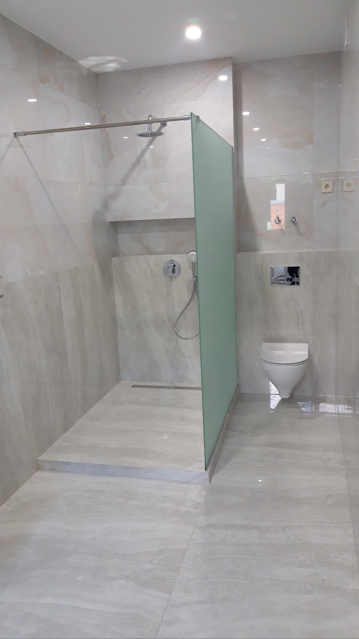 Nowoczesna łazienka z kabiną typu walk-in i beżowymi płytkami imitującymi marmur. Widoczne elementy armatury i podwieszana toaleta. Minimalistyczny design.