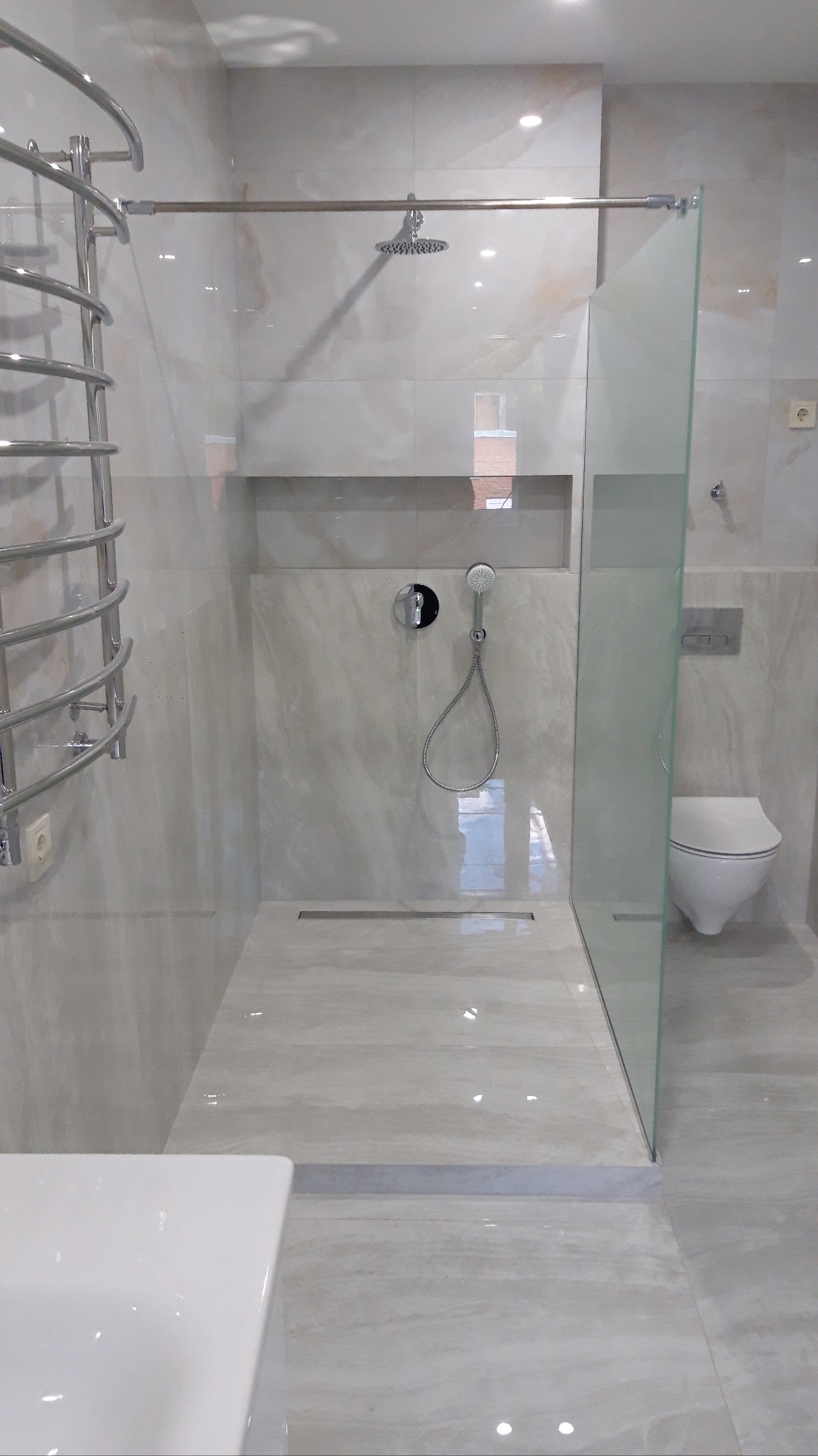 Stylowa kabina walk-in z odpływem liniowym, wykończona płytkami imitującymi marmur. Chromowana armatura i grzejnik drabinkowy, minimalistyczna toaleta podwieszana.