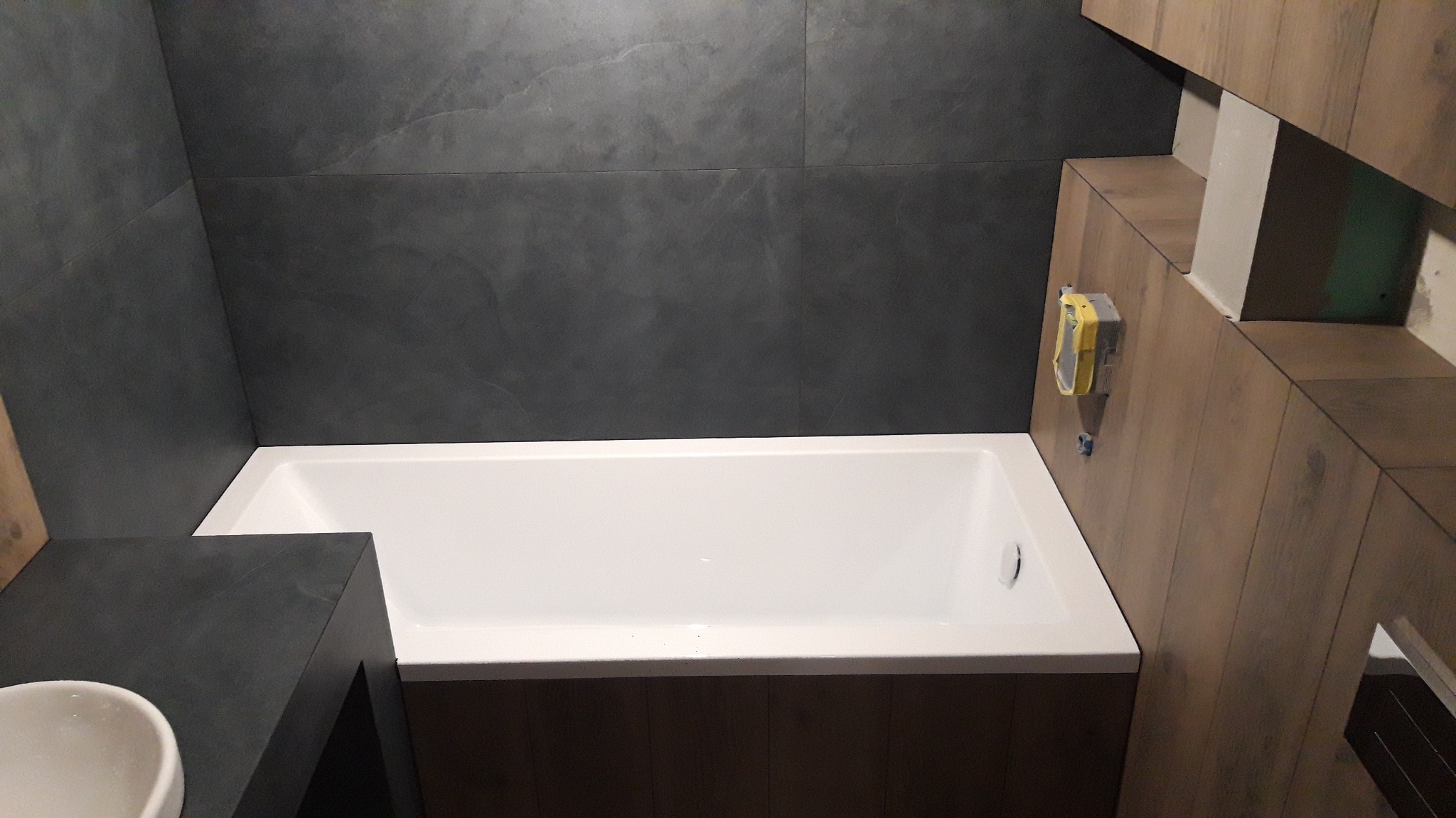 Nowoczesna łazienka z wanną obudowaną drewnopodobnymi panelami, szare płytki na ścianie i element umywalki. Widoczny fragment zabudowy z instalacją.